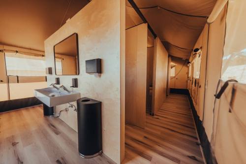 Interior of a modern tent bathroom with sinks, mirrors, and private stalls, featuring wooden flooring and natural light.