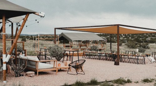 Outdoor seating area with wooden furniture, shaded tents, and desert landscape under a cloudy sky.