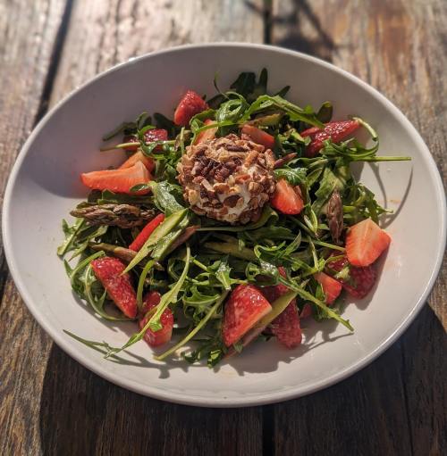 A fresh salad with arugula, strawberries, asparagus, and a round cheese ball, served in a white bowl on a wooden table.