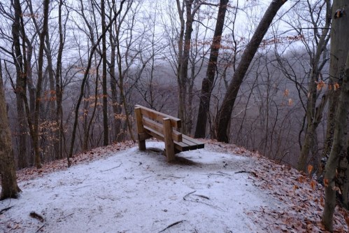 A wooden bench on a snowy hillside surrounded by bare trees and a misty landscape in the background.