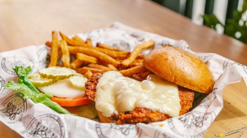 A crispy chicken sandwich with melted cheese, lettuce, tomato, pickles, and a side of golden fries on a paper-lined plate.