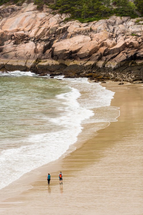 Two people walk along a sandy beach, with rocky cliffs and gentle waves in the background.