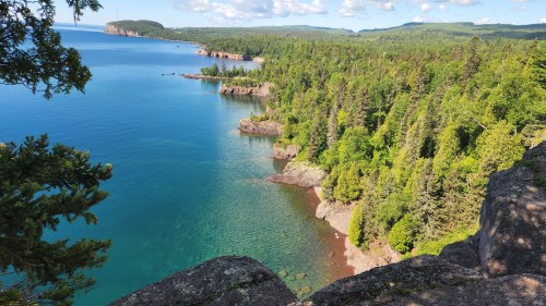 A scenic view of a calm blue lake surrounded by lush green forests and rocky shores under a clear sky.