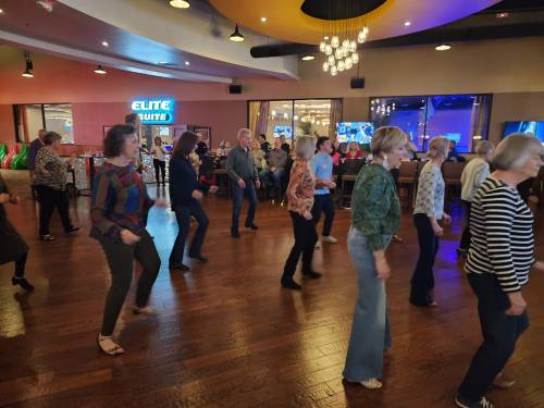 A group of people line dancing in a lively venue with colorful lights and a festive atmosphere.