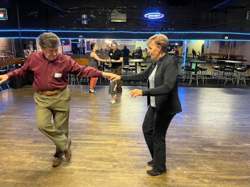 Two people dance on a wooden floor in a lively venue, with others enjoying the atmosphere in the background.