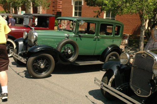 A vintage green car parked beside another classic car at a car show, with people and trees in the background.