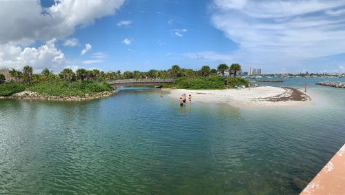 A scenic view of a calm lagoon with a sandy beach, palm trees, and people enjoying the water under a blue sky.