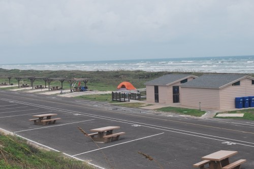 A coastal campsite with picnic tables, an orange tent, and beach access under a cloudy sky.