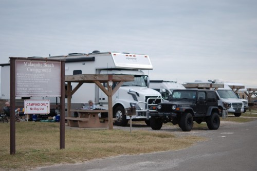 Sign for Malaguite Beach Campground with RVs and a black Jeep parked nearby on a cloudy day.