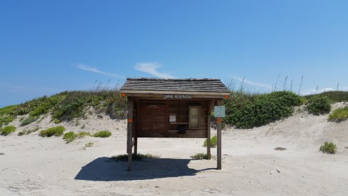 A wooden camping registration sign on a sandy beach, surrounded by green vegetation and under a clear blue sky.