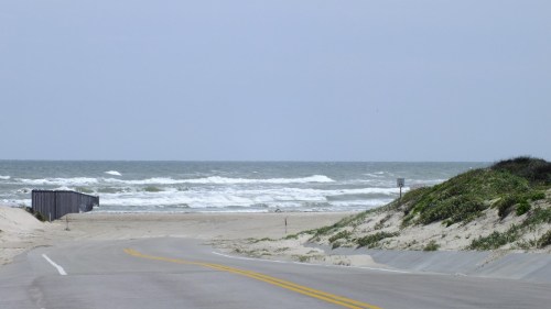 A winding road leads to a beach with waves crashing, bordered by a fence and coastal vegetation under a cloudy sky.