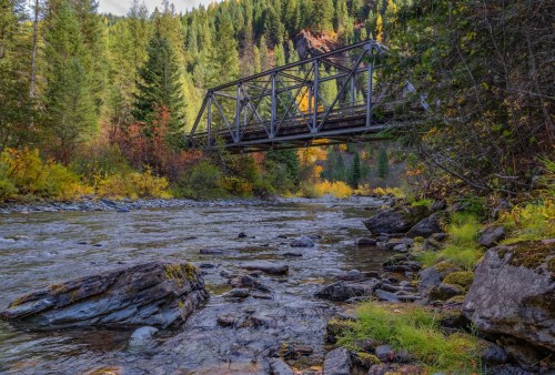 A metal bridge spans a river surrounded by lush green trees and rocky banks, with autumn colors in the foliage.