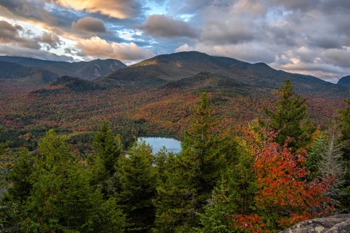 A scenic view of a colorful autumn landscape with mountains, a lake, and vibrant foliage under a cloudy sky.
