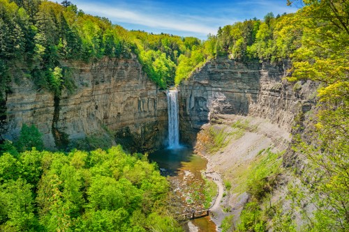 A stunning waterfall cascades into a lush green gorge, surrounded by trees and rocky cliffs under a clear blue sky.