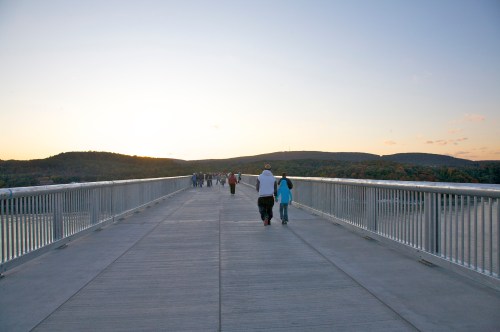 A wide pedestrian bridge at sunset, with people walking towards the horizon and hills in the background.