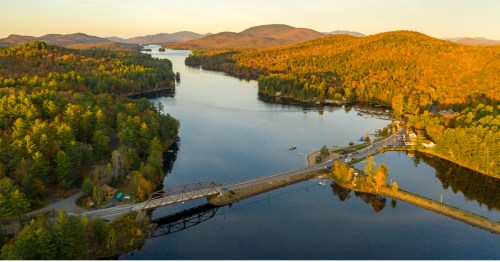 Aerial view of a serene lake surrounded by autumn-colored mountains and trees, with a bridge and small town visible.