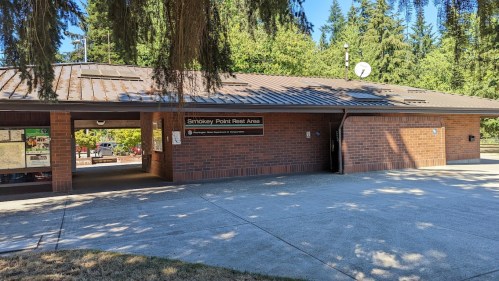 A brick building with a metal roof, labeled "Smokey Point Rest Area," surrounded by trees and a paved area.