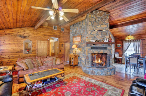 Cozy wooden living room with a stone fireplace, plush sofas, and a warm atmosphere, featuring rustic decor and a dining area.