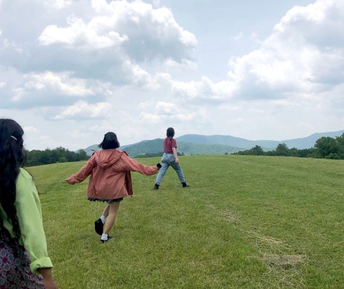 Three people walk across a grassy field with mountains in the background under a partly cloudy sky.