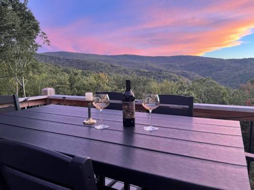 A table set with two wine glasses and a bottle, overlooking a scenic sunset view of mountains and trees.