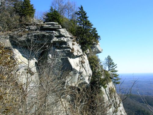 Rocky cliffside with sparse trees against a clear blue sky, overlooking a distant landscape.