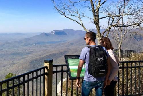 A couple stands at a viewpoint, looking out over a scenic mountain landscape with a sign nearby.