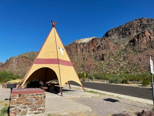 A colorful teepee structure near a road, set against a backdrop of rocky mountains and clear blue sky.