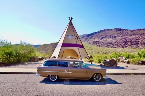 A vintage car parked beside a large teepee structure against a backdrop of mountains and clear blue sky.
