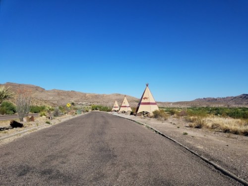 A deserted road leads to three teepee structures against a clear blue sky and desert landscape.