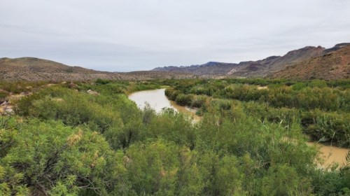A winding river flows through lush greenery, surrounded by rugged hills under a cloudy sky.
