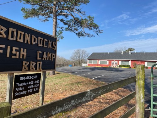 Sign for Boondocks Fish Camp BBQ with a building in the background, surrounded by trees and a clear blue sky.