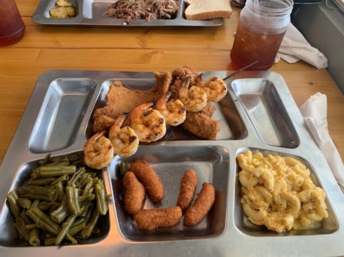 A metal tray with fried chicken, shrimp skewers, green beans, macaroni and cheese, and hushpuppies, alongside a drink.