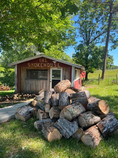 A rustic smokehouse with a sign, surrounded by green trees and a stacked pile of logs in the foreground.
