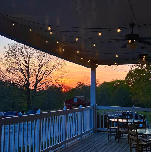 A sunset view from a porch with string lights, surrounded by trees and a glimpse of a red building in the distance.