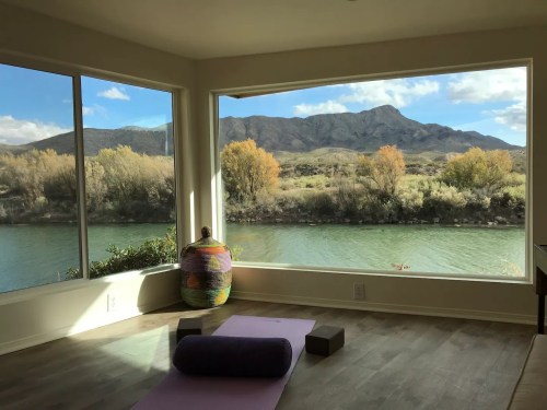 A serene room with large windows overlooking a river and mountains, featuring a yoga mat and cushions on the floor.