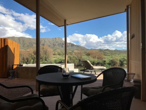 A scenic view from a patio featuring mountains, clouds, and greenery, with a table and chairs in the foreground.