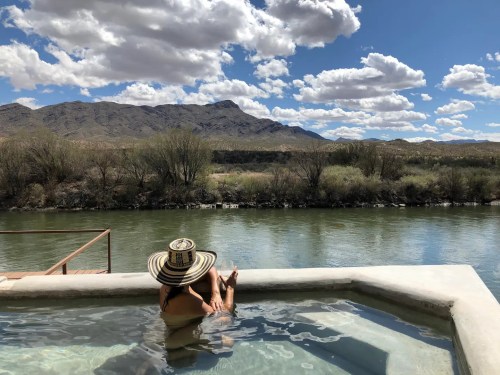 A person relaxes in a hot tub by a river, surrounded by mountains and a blue sky with fluffy clouds.