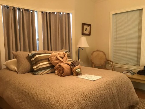 Cozy bedroom with a neatly made bed, decorative pillows, and soft blankets, featuring curtains and a lamp.