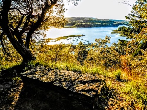 A scenic view of a lake surrounded by trees, with a stone bench in the foreground and sunlight reflecting on the water.