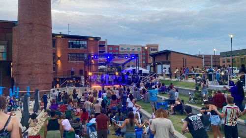 A lively outdoor concert scene with a crowd enjoying music under blue lights, surrounded by buildings and a cloudy sky.