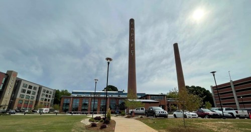 Historic industrial building with two tall smokestacks, surrounded by greenery and parked cars under a cloudy sky.
