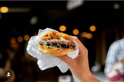 A hand holding a delicious burger with melted cheese and toppings, set against a blurred background.