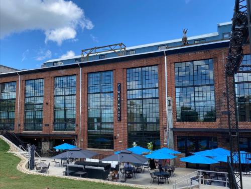 A brick building with large windows, featuring outdoor seating under blue umbrellas and a clear blue sky.