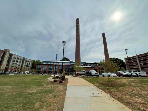 A modern building with two tall smokestacks, surrounded by a grassy area and parked cars under a cloudy sky.