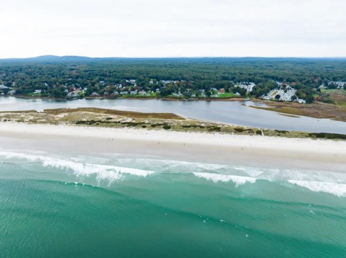 Aerial view of a beach with gentle waves, surrounded by greenery and a small coastal town in the background.