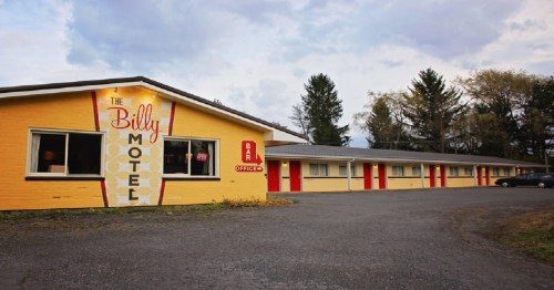 A yellow motel building with red doors and a sign reading "The Billy Motel" under a cloudy sky.