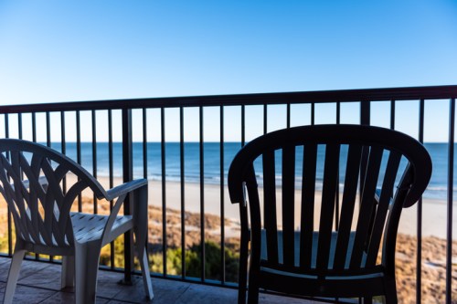 Two chairs on a balcony overlooking a calm beach and ocean under a clear blue sky.