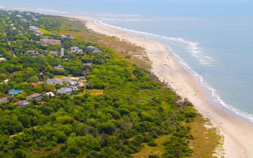 Aerial view of a sandy beach lined with greenery and houses, stretching along the coastline under a clear blue sky.