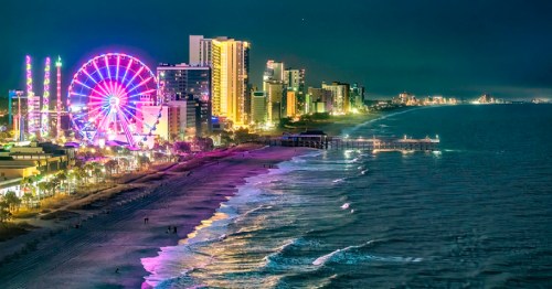 A vibrant night scene of a beachside city with a ferris wheel, illuminated buildings, and calm ocean waves.