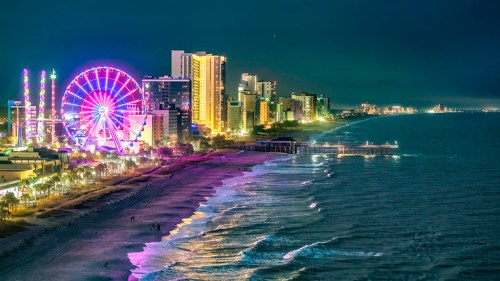 A vibrant city skyline at night with a ferris wheel, illuminated buildings, and a calm beach along the shoreline.